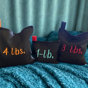 May include: Three black fabric weight bags with orange, teal, and red embroidered text. The bags are labeled 4 lbs. with an orange strap, 1-lb. with a teal strap, and 3 lbs. with a red strap. Each bag features a small fabric loop at the top.