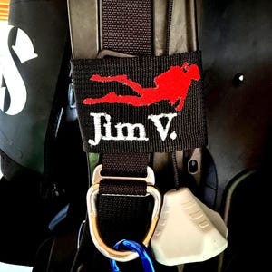 May include: A black fabric strap attached to scuba gear featuring an embroidered red silhouette of a scuba diver and the white text Jim V. The strap includes a silver metal D-ring, a blue carabiner, and a light gray plastic pull knob.