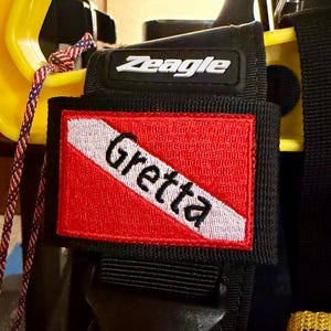 May include: A black Zeagle scuba diving equipment strap featuring a rectangular patch with a red and white diagonal stripe design. The name Gretta is embroidered in black thread across the white stripe on the fabric patch.