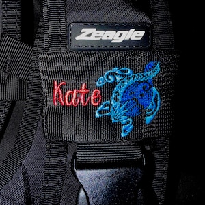 May include: A black Zeagle scuba diving buoyancy compensator strap featuring an embroidered patch. The patch displays the name Kate in red cursive thread next to a blue embroidered scorpion graphic. A black plastic buckle is visible below.
