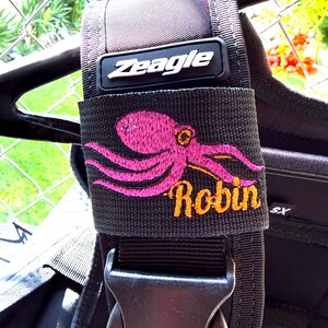May include: A black Zeagle scuba diving buoyancy compensator shoulder strap featuring an embroidered pink octopus graphic and the name Robin stitched in orange thread. The strap includes a black plastic side-release buckle and a rubberized logo.