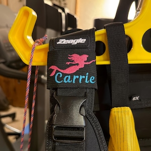 May include: A black Zeagle scuba diving buoyancy compensator shoulder strap featuring an embroidered pink mermaid silhouette above the name Carrie stitched in light blue thread. The strap includes a black plastic side-release buckle and webbing.