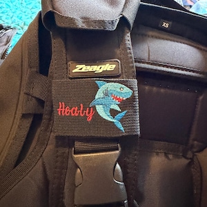 May include: A black Zeagle brand scuba diving buoyancy compensator vest featuring an XS size tag and a rectangular patch. The patch displays an embroidered blue shark graphic and the red cursive text Healy, positioned above a black plastic buckle.