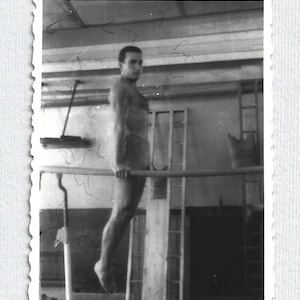 Può includere: Fotografia vintage in bianco e nero di una persona appesa a una sbarra orizzontale. La persona è senza maglietta e indossa pantaloncini. Lo sfondo comprende finestre e un muro.