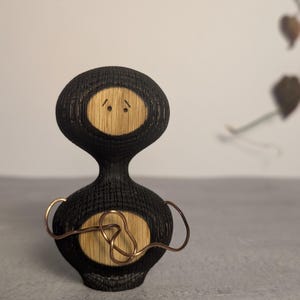 May include: A small, stylized wooden figurine with a black-stained exterior and natural wood-toned circular face and belly. The figure features a simple etched facial expression and a thin, curved copper-colored wire wrapped around its midsection.