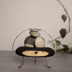 May include: A decorative object featuring a circular, charred wood base with a light-colored face, supported by three thin copper-colored metal legs. A thin copper-colored wire arch extends over the top, holding a stack of five smooth, grey river stones.