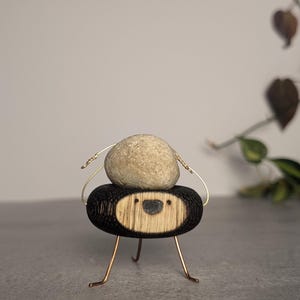 May include: A whimsical wooden sculpture of a creature with a light brown stone head, a black oval body with a light brown face, and thin copper wire legs and arms. The sculpture is on a gray surface.
