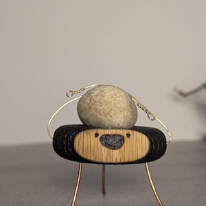 May include: A whimsical sculpture featuring a smooth, gray stone balanced on a wooden base with a smiling face design. The base is black with a natural wood front, supported by thin, copper-colored legs and wire accents.