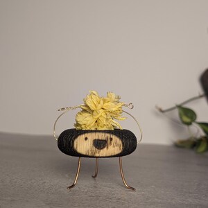 May include: A small, oval-shaped wooden figure with a charred black exterior and a light-colored face featuring a simple burned-in smile. The figure has thin, gold-toned wire legs and a wire loop on top holding a dried, pale yellow flower cluster.