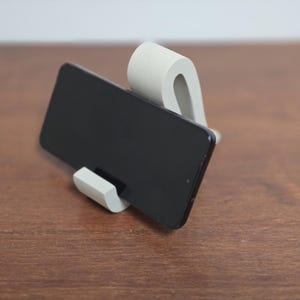 Peut inclure: Un support de téléphone en béton gris tenant un smartphone noir. Le support a un design incurvé avec un petit rebord pour soutenir le téléphone. Le téléphone est positionné horizontalement sur une surface en bois.