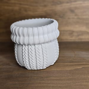 May include: A light grey ceramic planter with a textured, sweater-style design. The top has a ribbed collar, and the body features a cable knit pattern. The planter sits on a wooden surface.