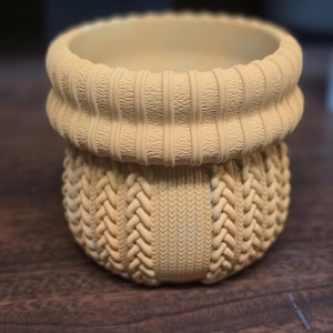 May include: A beige ceramic plant pot with a textured knit design. The top rim has a ribbed pattern, while the body features a braided knit pattern. The pot is designed to hold small plants or succulents.