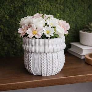 May include: A grey ceramic flower pot with a knitted sweater design, filled with pink and white flowers. The pot is on a dark wooden shelf, with books and a candle in the background.