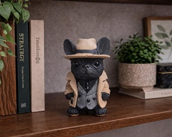 May include: A black bulldog figurine wearing a tan fedora, trench coat, and a gray vest and tie. The dog statue is standing on a wooden shelf. A small potted plant, books, and a framed picture are in the background.