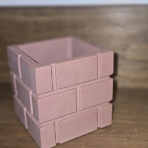 Puede incluir: Una maceta cuadrada rosa con un diseño similar a ladrillos. La maceta está hecha de un material sólido y tiene una abertura rectangular en la parte superior. El color es un rosa apagado y polvoriento.