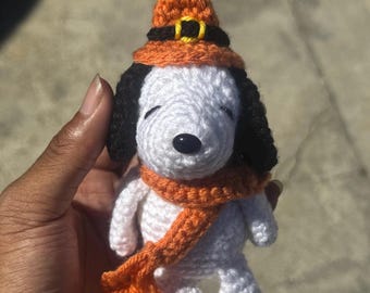 May include: Crocheted Snoopy figure wearing an orange witch hat with a black band and yellow buckle, and an orange scarf. The dog has black ears and a black nose. The figure is white with black accents.