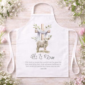 May include: White apron with a watercolour illustration of a lamb, a wooden cross, and olive branches. The text "His Is Risen" and a Bible verse are printed below. The apron has white straps.