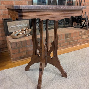 May include: A brown wooden side table with a rectangular marble top. The table has ornate carved details on the legs and apron. The table is standing on a light-colored rug.