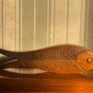 May include: A wooden fish sculpture with a detailed scale pattern. The fish is carved from a light brown wood and has a dark brown accent on the scales. The sculpture has a tail and fins, and is displayed on a wooden surface.