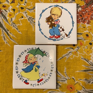 May include: Two ceramic tiles with colorful illustrations of children. The first tile features a boy holding a teddy bear and a dog. The second tile features a girl holding an umbrella and a dog. Both tiles have a white background with blue and green accents.