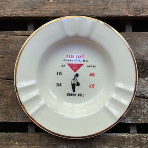 May include: White ceramic ashtray with a gold rim and a black and white illustration of a bowler. The ashtray has the text "Park Lanes Charlotte, N.C. 32 Lanes 215 600 240 635 Honor Roll" printed on it.