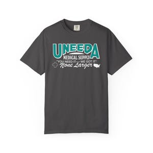 May include: Dark gray t-shirt with the text "UNEEDA MEDICAL SUPPLY" in teal and white. The shirt also features the phrases "YOU NEED IT--WE GOT IT" and "None Larger" with a cross and a map of the United States.