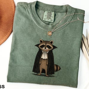 May include: A moss green short-sleeve t-shirt featuring a graphic of a raccoon dressed as a vampire in a black cape and white cravat. The shirt has a crew neckline and a Comfort Colors brand label printed on the inside neck area with size S.