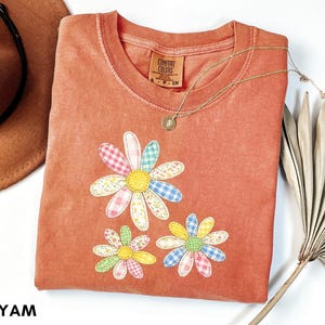 Puede incluir: Una camiseta Comfort Colors de color coral con tres aplicaciones de margaritas en patrones de cuadros pastel. Un collar de oro con un pequeño colgante descansa sobre la camisa. Un sombrero marrón y hojas de palma secas están en el fondo.