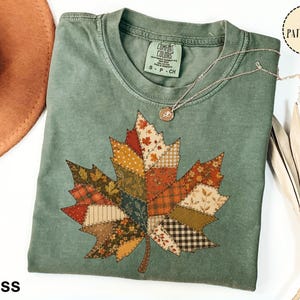 Puede incluir: Una camiseta de cuello redondo color verde musgo con un gráfico grande de hoja de arce compuesto por varios patrones de tela estilo patchwork en tonos naranja, marrón, crema y verde oscuro. La camiseta tiene una etiqueta Comfort Colors en el cuello.