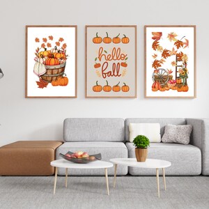Könnte beinhalten: Drei gerahmte Kunstdrucke zum Thema Herbst. Die Drucke zeigen Aquarellillustrationen von Kürbissen, Blättern und dem Text "hello fall". Das Kunstwerk wird über einem grauen Sofa und einem Couchtisch ausgestellt.