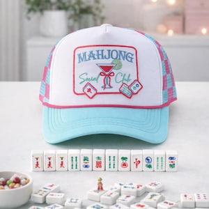 May include: A white and blue Mahjong Social Club trucker hat with a pink checkered mesh back, and a light blue brim. The hat features an embroidered logo with a cocktail glass and dice. Mahjong tiles are arranged in front of the hat.