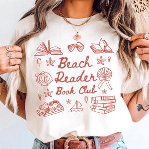 Può includere: T-shirt color crema con la scritta "Beach Reader Book Club" in rosso, insieme a illustrazioni di libri, conchiglie e altri elementi a tema spiaggia. La persona indossa jeans blu e una cintura marrone con fibbia decorativa.