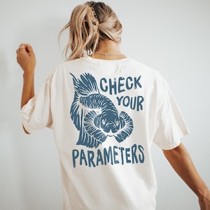 Könnte beinhalten: Cremefarbenes T-Shirt mit einer blauen Illustration eines Fisches und dem Text "CHECK YOUR PARAMETERS". Das Grafik-T-Shirt hat eine lockere Passform und einen Rundhalsausschnitt. Das Design ist auf der Rückseite des Shirts aufgedruckt.