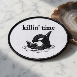 May include: Oval patch with a black border and a white center. The patch features a black orca reading a book in water, with the words "killin' time" above. The orca is stylized and the text is in a simple font.