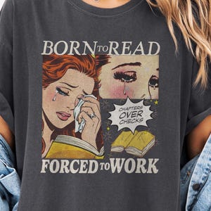May include: Dark gray t-shirt with a comic-style graphic. The graphic features a crying woman holding a book, with text that reads "BORN TO READ" and "FORCED TO WORK." A speech bubble says "CHAPTERS OVER CHECKS."