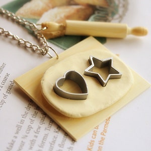 May include: A wooden pendant necklace with a silver chain. The pendant features a small rolling pin and a piece of white clay with a heart and star cookie cutter on top.