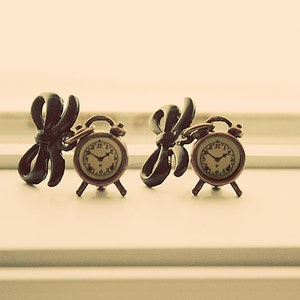 May include: A pair of black bow earrings with small gold alarm clock charms. The clocks have black hands and Roman numeral markings.