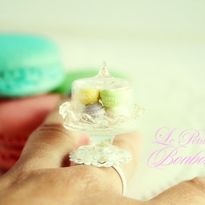Könnte beinhalten: Ein transparenter Glasring mit einer weißen Basis und einer Glaskuppel, gefüllt mit bunten Macarons. Der Ring befindet sich an einer Hand und der Text "Le Petite Bombon" ist im Hintergrund.