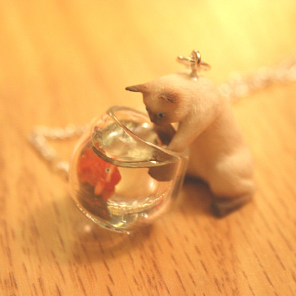 Mischievous Siamese Kitty cat is playing with goldfish in a fish bowl necklace