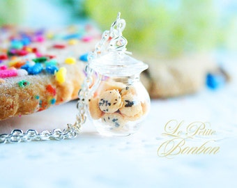 May include: A silver necklace with a clear glass jar charm filled with miniature chocolate chip cookies. The necklace is on a white surface with a blurred background of a cookie with colorful sprinkles.