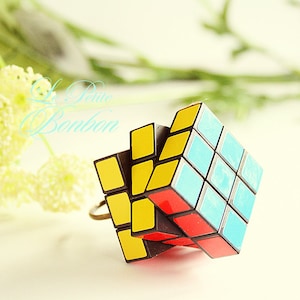May include: A Rubik's Cube ring with a partially solved puzzle design. The cube features yellow, blue, and red squares. The ring is set against a soft background with the text "La Petite Bonbon".