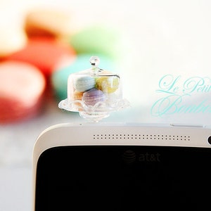 May include: A white smartphone with a black screen and the text "at&t" on the bottom. A miniature glass cake stand with a clear lid sits on top of the phone. The cake stand is filled with pastel-colored macarons. The text "Le Petite Boutique" is written in a light blue cursive font on the background.