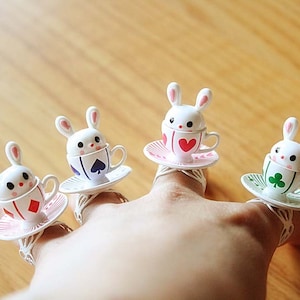 May include: Four white rabbit finger rings with teacups and saucers. Each ring features a different playing card symbol: a heart, a spade, a diamond, and a clover.
