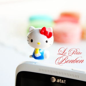 May include: A white Hello Kitty figurine with a red bow, blue dress, and yellow scarf, sitting on a white smartphone with the text "Le Petite Bonbon" and "at&t" on the screen.