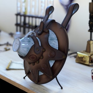 May include: A brown leather decorative piece with a circular design and star-shaped cutouts, holding a small, marbled glass cup. The leather has decorative stitching and rivets. The piece has two handles and stands on three black metal legs.