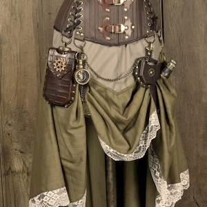 Może przedstawiać: Brązowy gorset w stylu steampunk ze złotymi klamrami i dopasowaną spódnicą z koronkową lamówką. Spódnica ma brązową skórzaną kieszeń z wzorem koła zębatego i zegarek kieszonkowy przymocowany do łańcuszka.