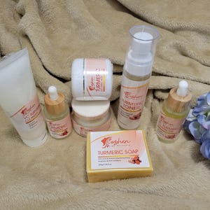 May include: A set of turmeric skincare products from the brand Coshen Pink by Nature. The set includes a cleanser, toner, serum, soap, and a face cream. The products are arranged on a light brown fuzzy surface with a blue flower in the background.