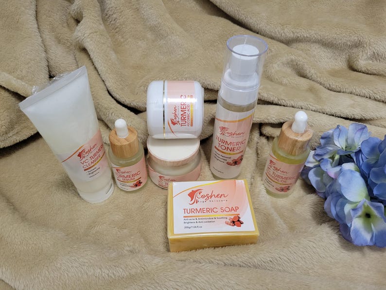 May include: A set of turmeric skincare products from the brand Coshen Pink by Nature. The set includes a cleanser, toner, serum, soap, and a face cream. The products are arranged on a light brown fuzzy surface with a blue flower in the background.