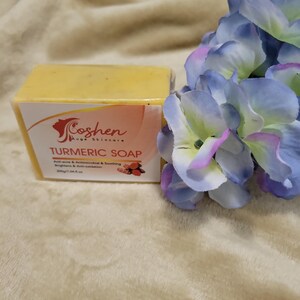 May include: A yellow bar of turmeric soap with the text "Coshen Ave Skincare Turmeric Soap" and "Anti-acne & Anti-microbial & Soothing Brightens & Anti-inflammation 200g/7 oz e" printed on the label. The soap is sitting on a light brown fuzzy surface with a blue flower in the background.