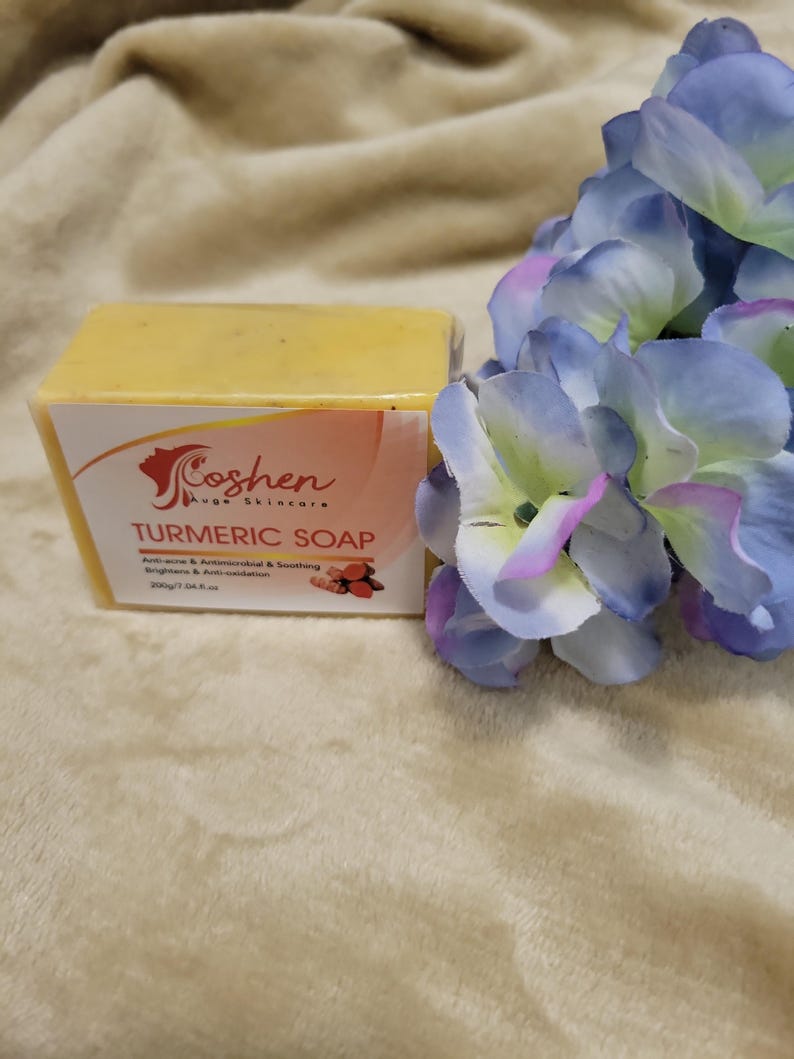 May include: A yellow bar of turmeric soap with the text "Coshen Ave Skincare Turmeric Soap" and "Anti-acne & Anti-microbial & Soothing Brightens & Anti-inflammation 200g/7 oz e" printed on the label. The soap is sitting on a light brown fuzzy surface with a blue flower in the background.